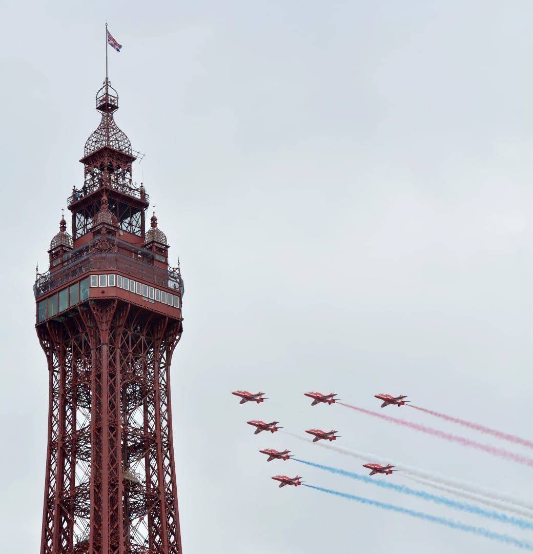 top of tower with 8 aeroplanes flying towards the tower with blue and red smoke billowing behind